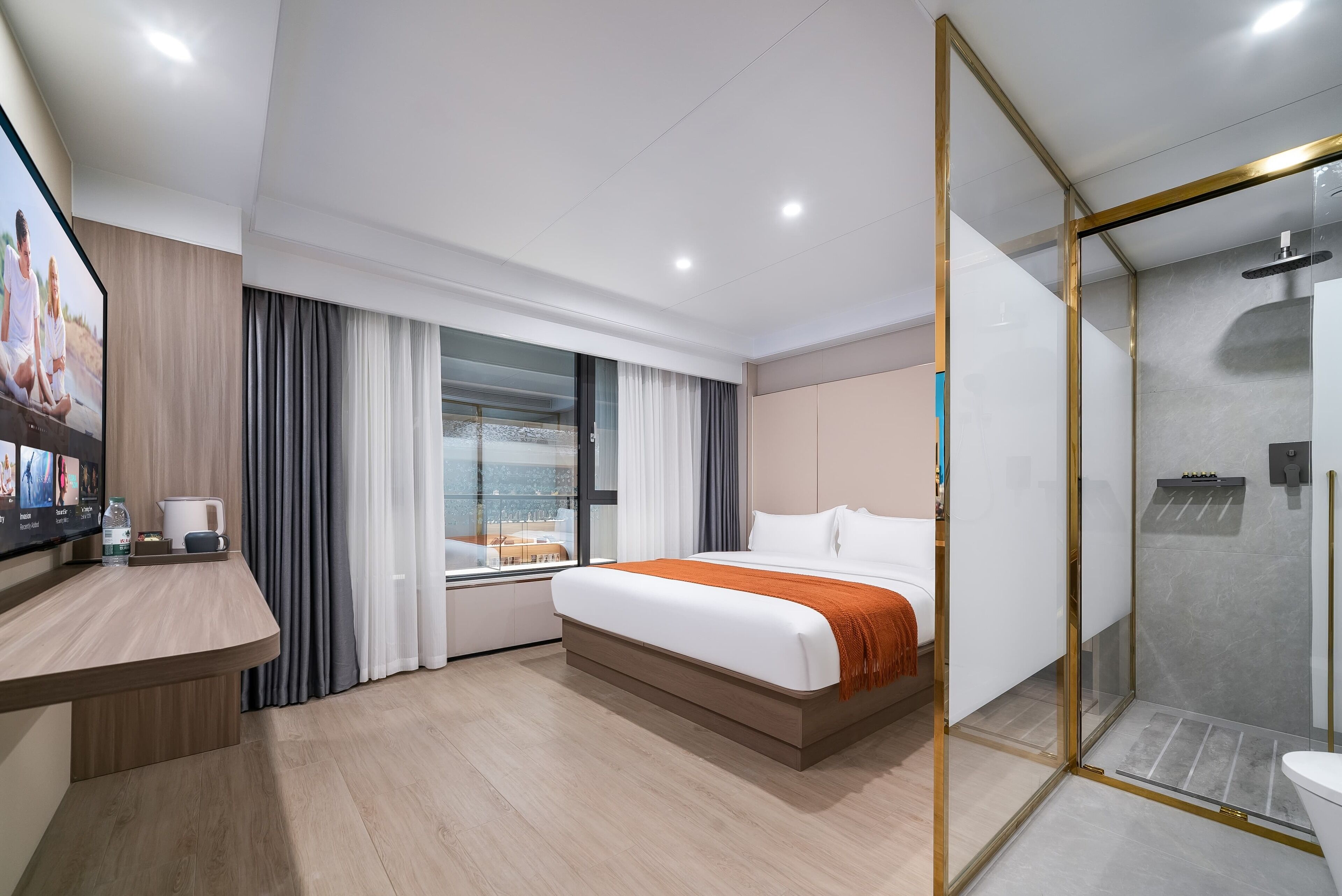 A modern hotel room with a large bed, a desk with a flat-screen TV, a glass shower enclosure, and a view of the city through large windows.