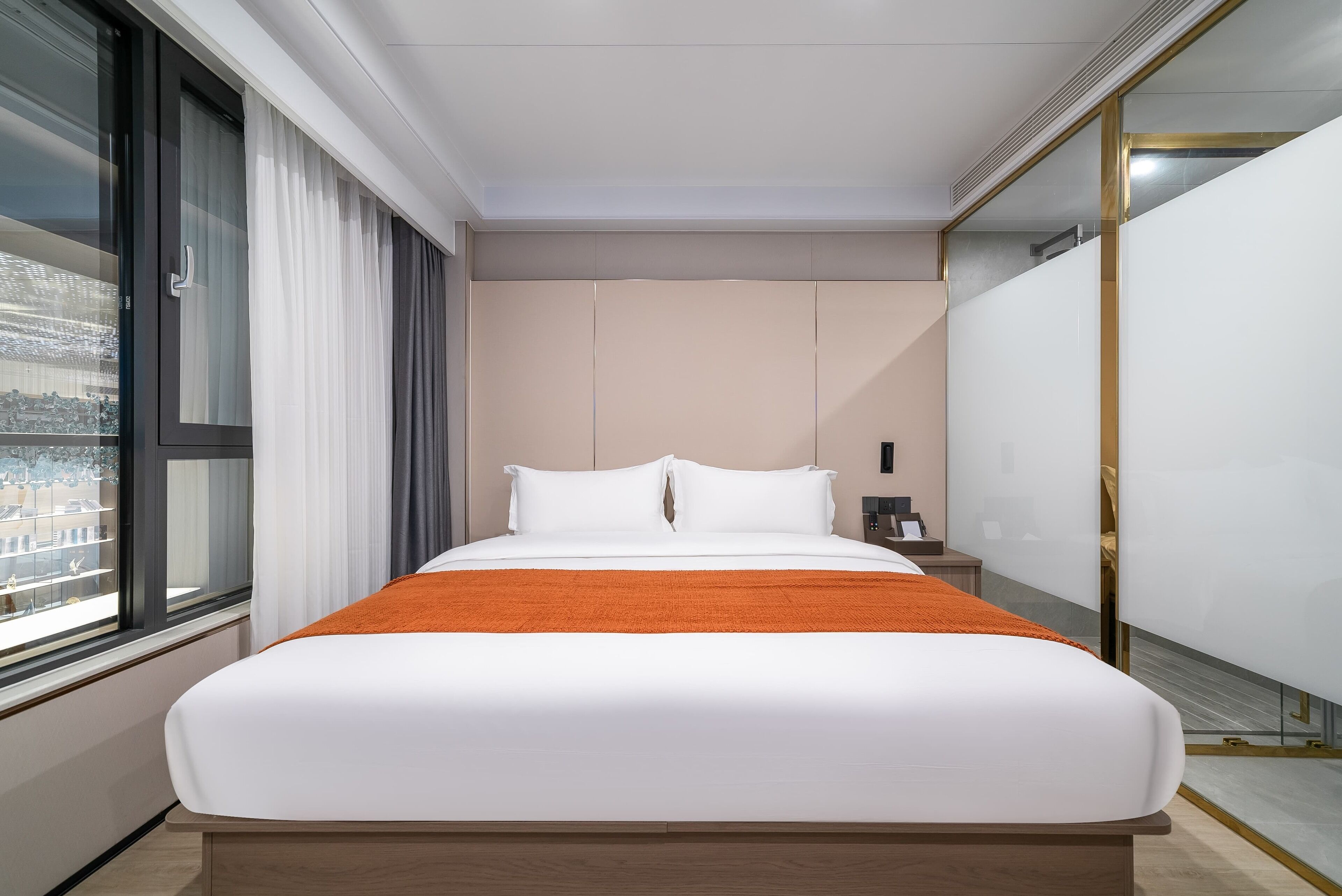 A modern hotel room with a large bed, a view through a window, and a bedside table with a lamp.