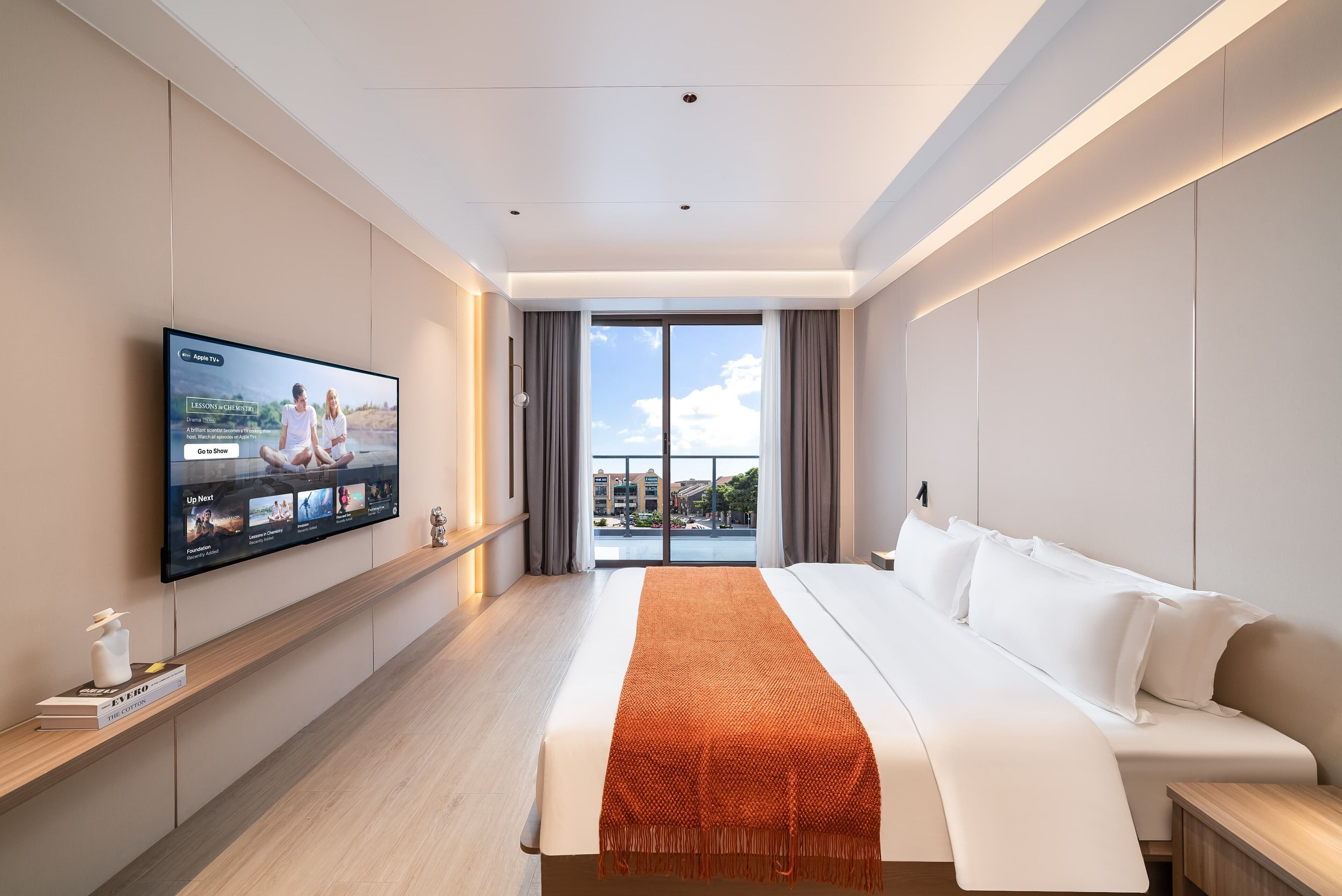 A modern hotel room with a large bed, a flat-screen TV mounted on the wall, a wooden bedside table, and a view of the city through the large window.