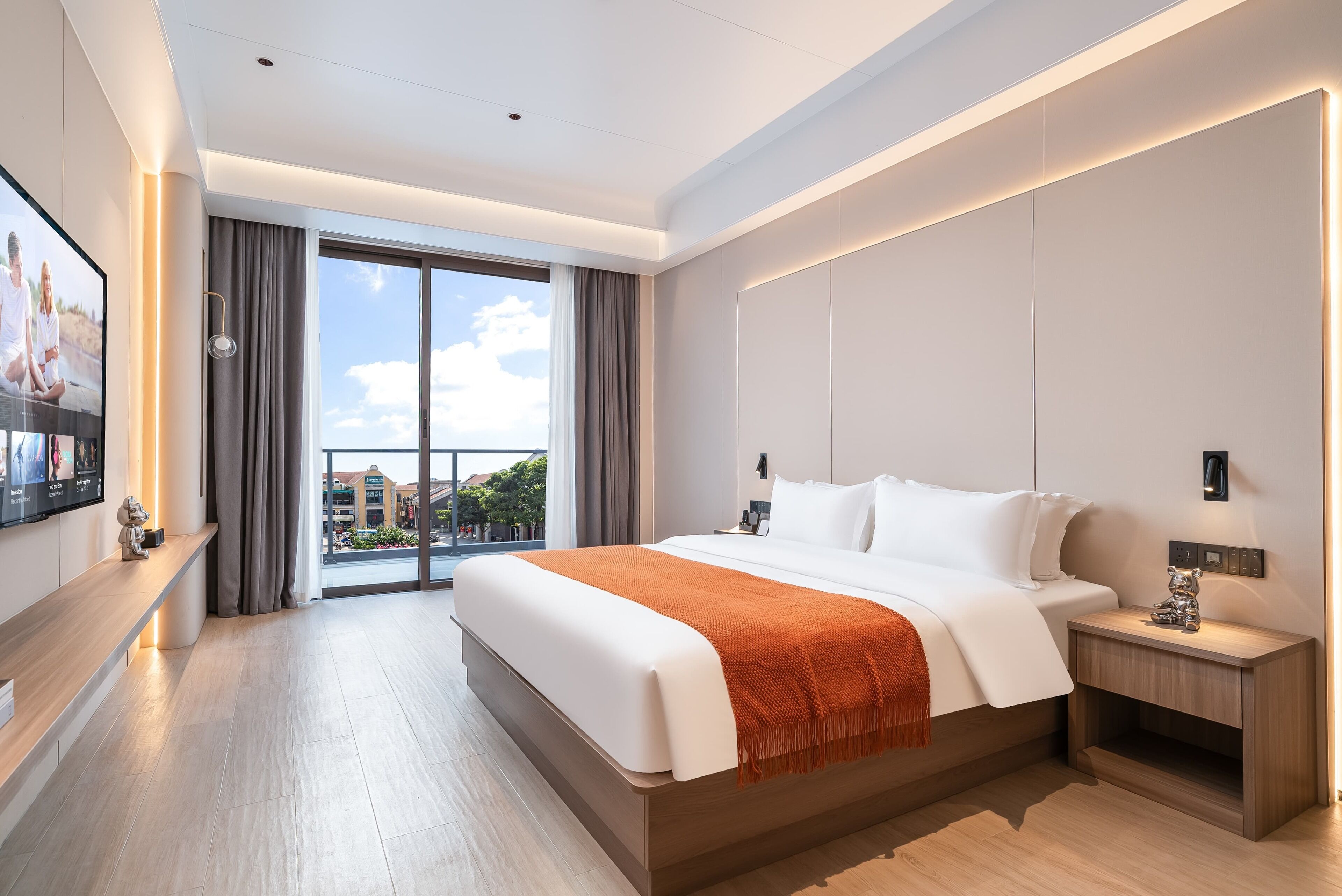 Habitación de hotel moderna con una cama grande, una mesita de noche, un televisor montado en la pared y vistas a la ciudad a través de amplios ventanales.