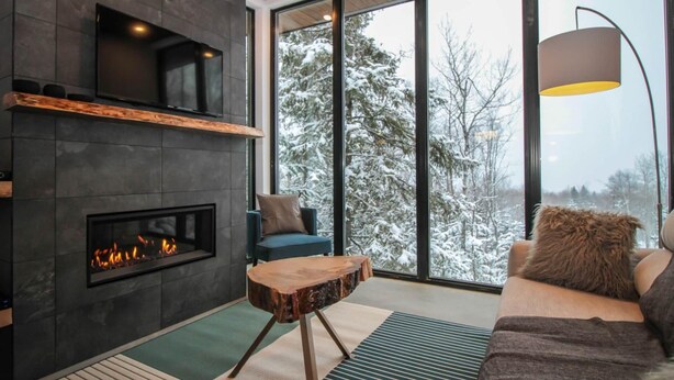 Ett modernt vardagsrum med en öppen spis, en soffa, ett soffbord och ett stort fönster med utsikt över ett snötäckt landskap.