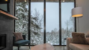 Ett modernt vardagsrum med ett stort fönster, ett soffbord i trä, en soffa och en lampa.