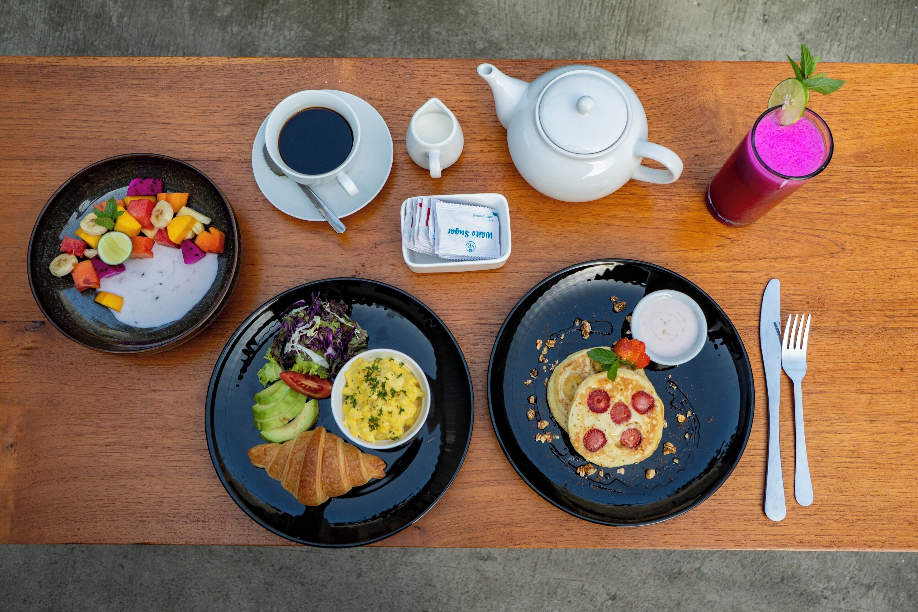 Ein Frühstückstisch mit verschiedenen Speisen, darunter Obstsalat, Rührei, Pfannkuchen und ein Croissant.