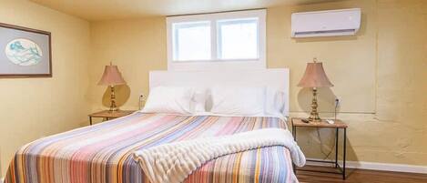 A bedroom with a bed, two bedside lamps, a window, an air conditioner, and a framed picture on the wall.