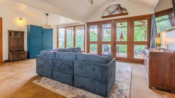 A spacious living room with a large sectional sofa, a ceiling fan, and a view through large windows. - The Bear’s Den at Triple Z (Medicine Park)