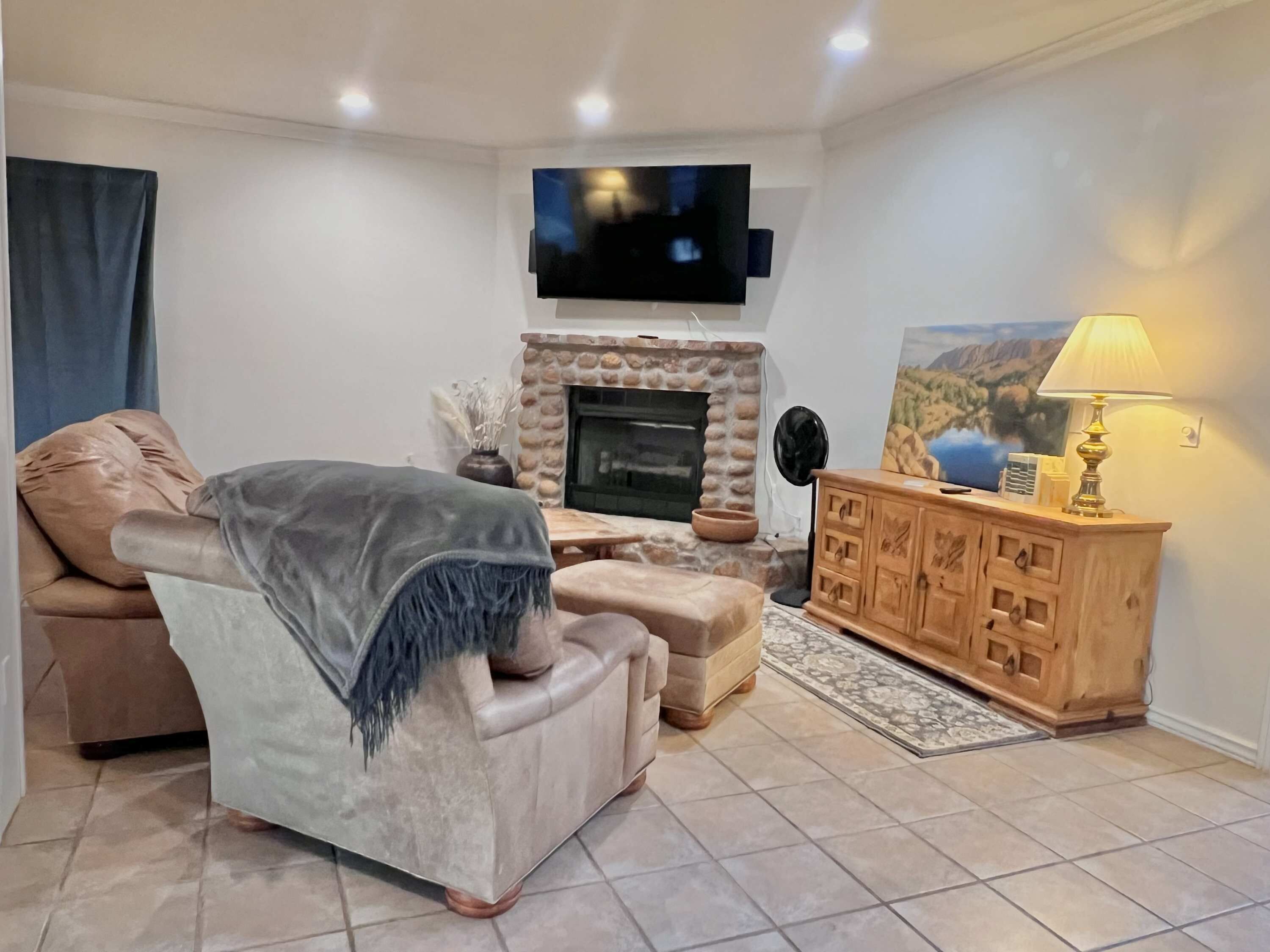 Un salon avec un canapé beige, un téléviseur fixé au mur, une cheminée et un meuble en bois sur lequel est posée une lampe.