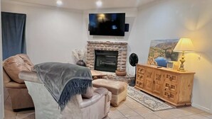 Un salon avec un canapé beige, un téléviseur fixé au mur, une cheminée et un meuble en bois sur lequel est posée une lampe.