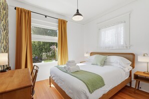 A neatly made bed with white linens and a green throw, a wooden nightstand with a lamp, a dining table with chairs, and a window with curtains. - Walk to Restaurants & Coffee + BBQ + Tesla/J1772 L1 EV Charger (Seattle)