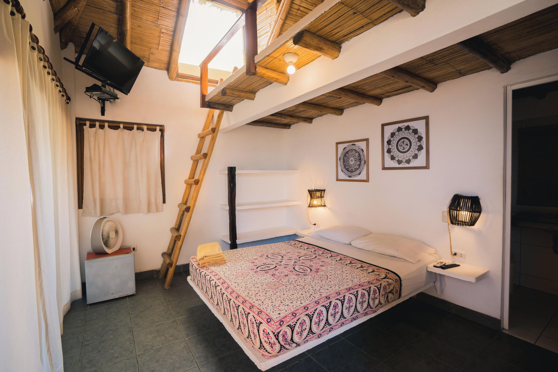 Un dormitorio con cama, mesitas de noche, un ventilador, una escalera, televisión y ventilador de techo.