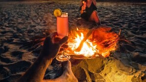 A beach scene with a fire, palm trees, and a building in the background. - Peña Linda Bungalows (Los Organos)
