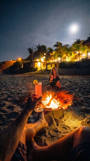Cena de praia com fogueira, palmeiras e um edifício ao fundo.