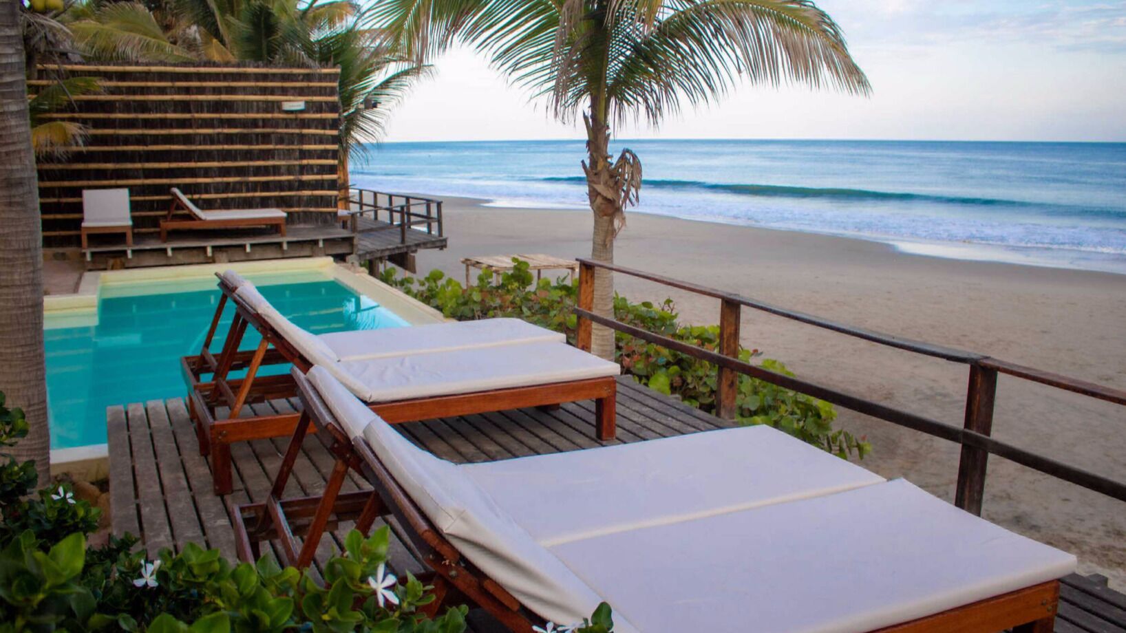 Zona de piscina con sillones de descanso y una terraza de madera con vista a una playa.