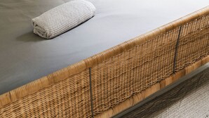 Una cama con colchón gris, cabecero tejido y mesita de noche con lámpara.