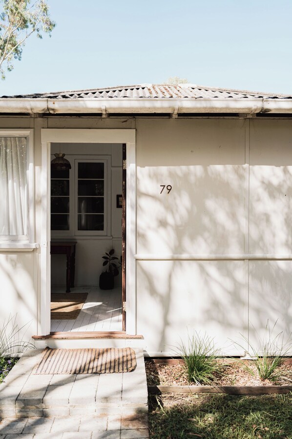 Un bâtiment blanc avec le chiffre « 79 » peint sur le côté, une porte et un petit porche avec un paillasson et des plantes.