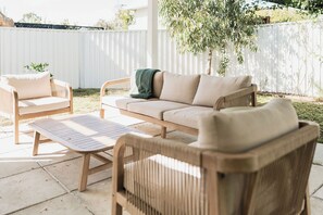 An outdoor seating area with wicker furniture, a cushioned bench, and a white fence in the background. - OB Surf Shack — relaxed, chic surf shack on the south coast (Denmark)