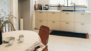 Una cocina con una mesa de comedor, sillas, una lámpara colgante, un ventanal con cortinas y una estantería con utensilios de cocina.