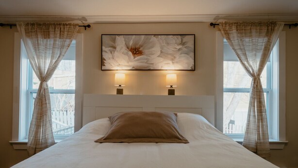 A neatly made bed with a large pillow, two bedside lamps, and a large floral painting above the bed. - Downtown Heritage Home | 3 Bed + Backyard (St. John`s)