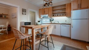A modern kitchen with wooden cabinets, a white refrigerator, a microwave, and a dining area with wicker chairs. - Downtown Heritage Home | 3 Bed + Backyard (St. John`s)