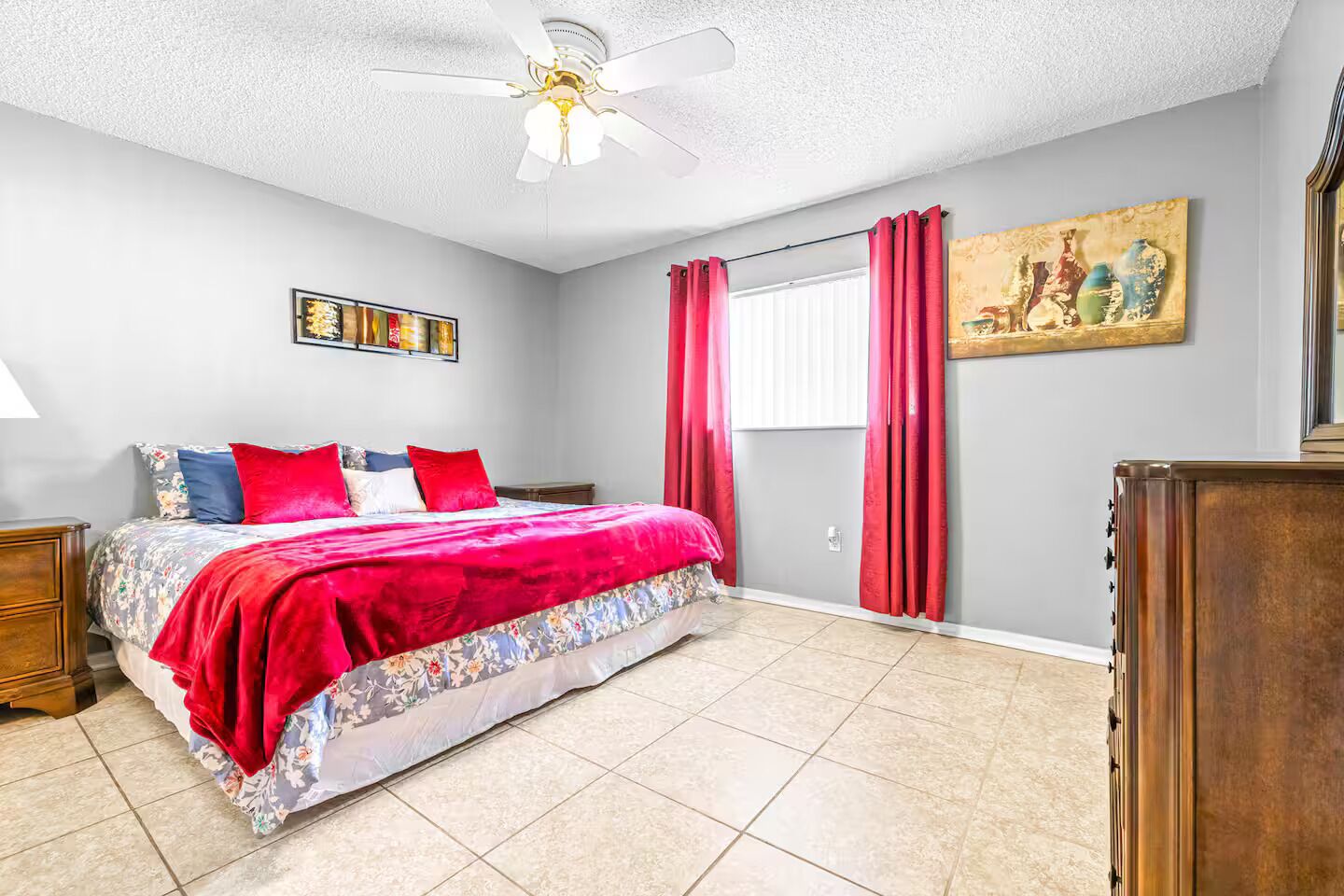 Un dormitorio con cama, ventilador de techo, ventana con cortinas, un cuadro en la pared y cómoda de madera.