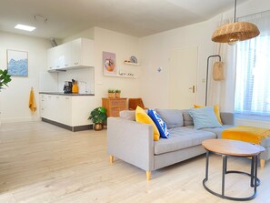 Ein modernes Wohnzimmer mit grauem Sofa, einem kleinen Holztisch und einer Küchenzeile mit weißen Schränken und einer schwarzen Arbeitsplatte.