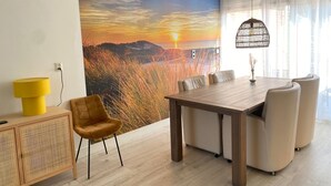 Een eethoek met een houten tafel en stoelen, een gele lamp, een rotan kast en een groot muurschildering van een zonsondergang landschap.