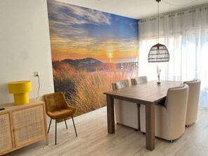 Ein Essbereich mit Holztisch und -stühlen, einer gelben Lampe, einem Rattankorpus und einem großflächigen Wandbild mit einer Sonnenlandschaft.