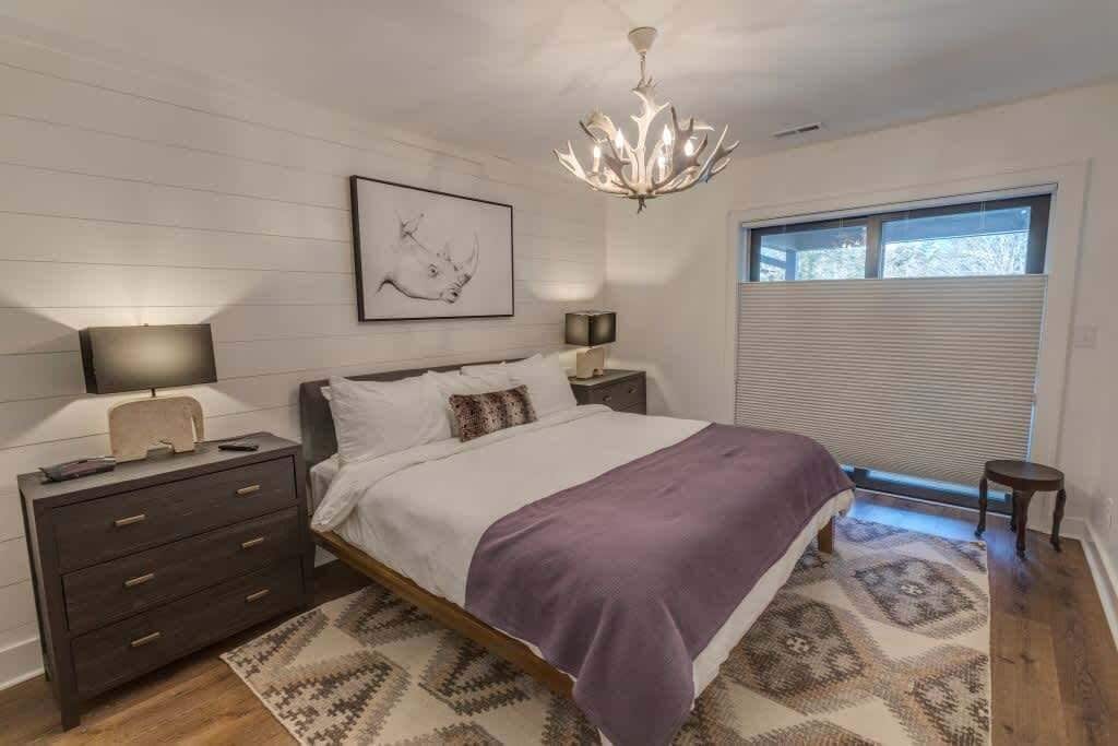 A bedroom with a bed, bedside tables, a chandelier, a window with blinds, and a rug.