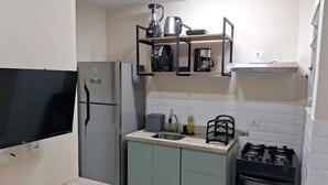 Eine kompakte Küche mit eingebautem Kühlschrank, einem Gasherd und einer Mikrowelle. Ein kleines Waschbecken und ein darüberliegendes Regal beherbergen eine Kaffeemaschine und einen Wasserkocher.