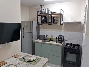 Eine kompakte Küche mit eingebautem Kühlschrank, einem Gasherd und einer Mikrowelle. Ein kleines Waschbecken und ein darüberliegendes Regal beherbergen eine Kaffeemaschine und einen Wasserkocher.