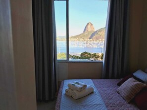 A hotel room with a bed, a window with curtains, and a view of a mountain and water. - Botafogo, Rio de Janeiro | Apartment with a view of Sugarloaf Mountain   (Rio de Janeiro)