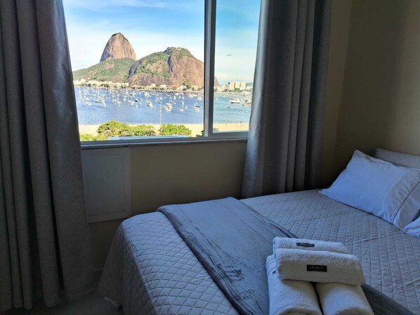 A hotel room with a bed, a window with curtains, and a view of a mountain and marina. - Botafogo, Rio de Janeiro | Apartment with a view of Sugarloaf Mountain   (Rio de Janeiro)