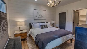 A bedroom with a bed, bedside tables, a chandelier, a door, and a wall-mounted artwork.