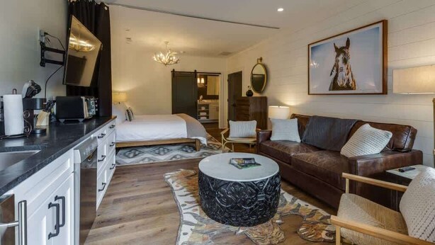 A modern hotel room with a bed, a sofa, a coffee table, a desk with a computer, and a dining area.