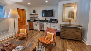 A modern kitchen with white cabinets, a microwave, and a coffee maker. There are two wooden chairs with cushions. A wooden dresser and a framed picture are on the wall.