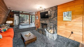 A cozy living room with a stone fireplace, a wooden wall, and a staircase leading to an outdoor area. - The Mod Lodge, Retro Sunriver Cabin Vibes! (Sunriver)