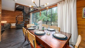 A dining area with a wooden table set for four, a spiral staircase, and large windows with curtains. - The Mod Lodge, Retro Sunriver Cabin Vibes! (Sunriver)