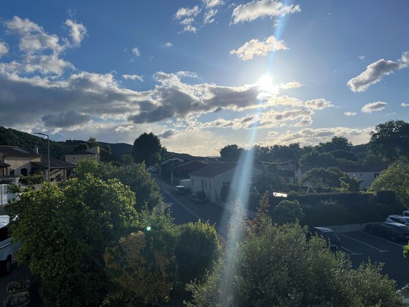 A residential area with houses, parked cars, and trees under a partly cloudy sky with the sun shining through.