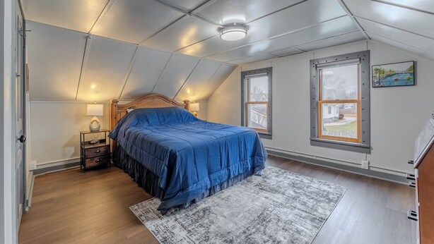 Et soveværelse med skråvæg, en seng med blåt sengetæppe, et natbord med lampe, og to vinduer med udsigt til et hus og en strand.