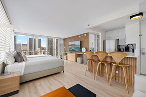 A modern hotel room with a large bed, a dining area with bar stools, a kitchen, and a city view. - Newly Innovated Oceanview Beach Condo Sauna Pool (Honolulu)