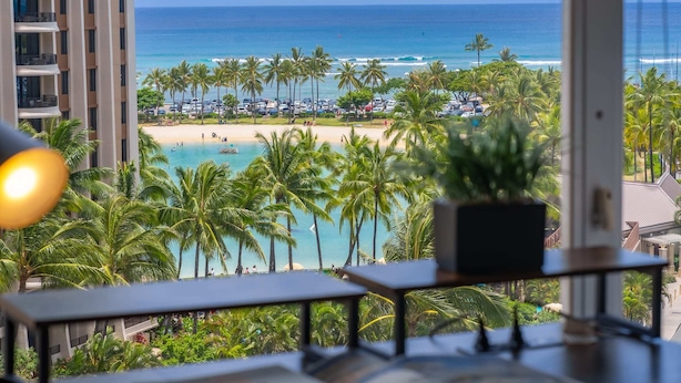 Blick vom Balkon auf einen Strand mit Palmen, ein Schwimmbad und das Meer.