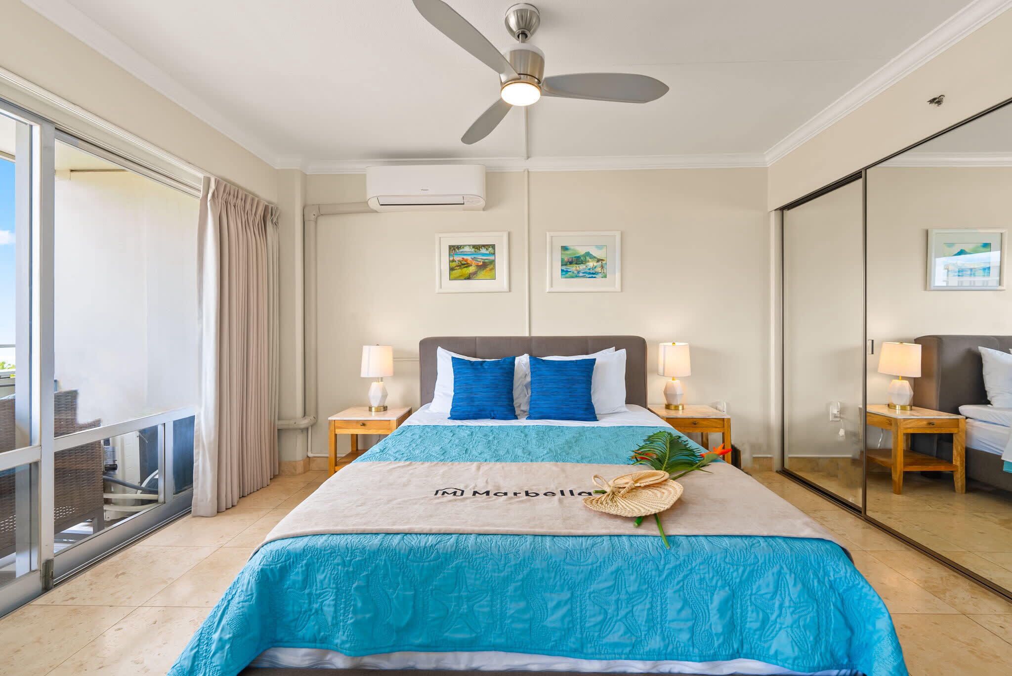 Quarto de hotel com uma cama, um ventilador de teto, duas mesas de cabeceira com luminárias, uma janela grande com cortinas e uma varanda.