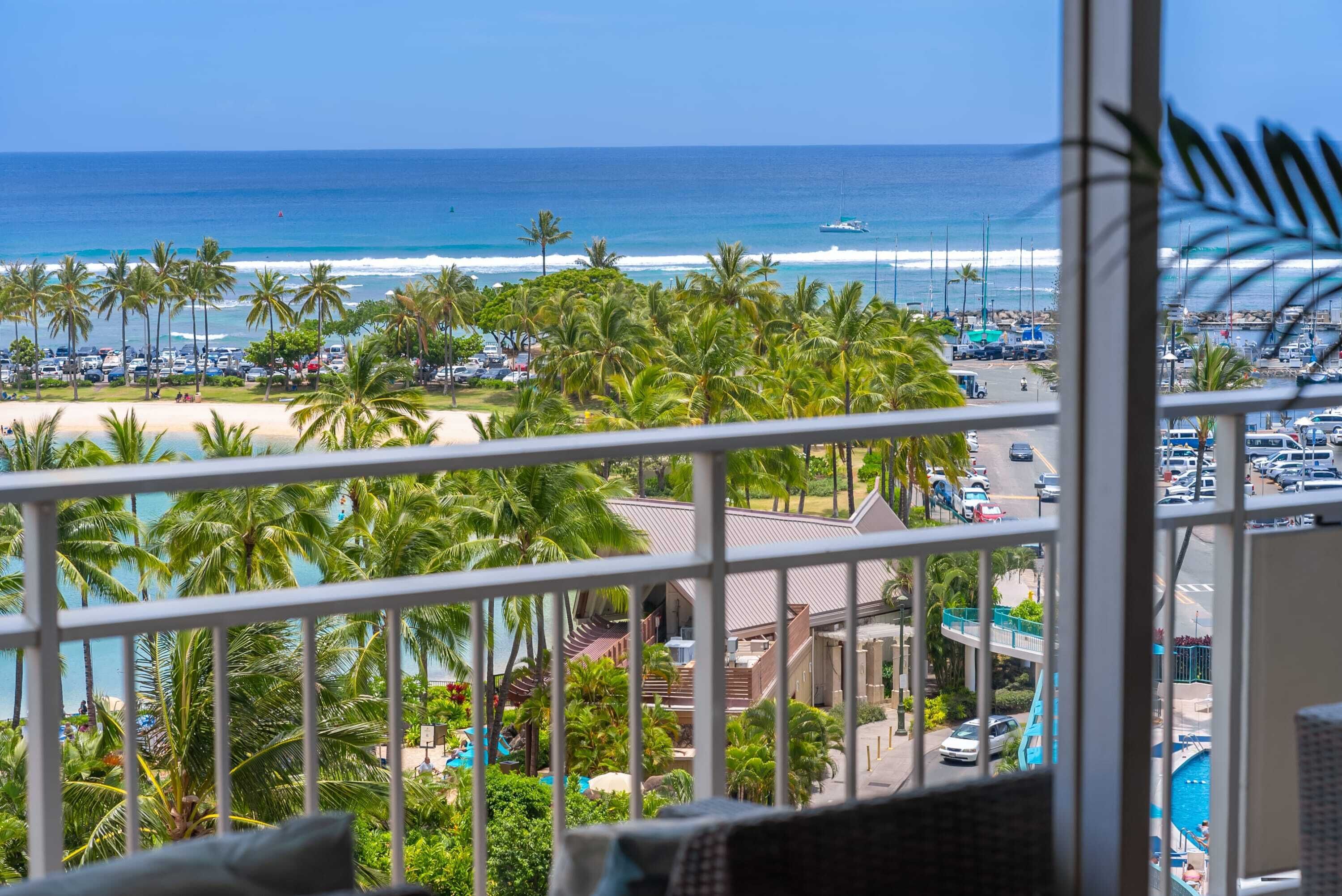 A view from a balcony overlooking a beach with palm trees, a pool, and a marina.