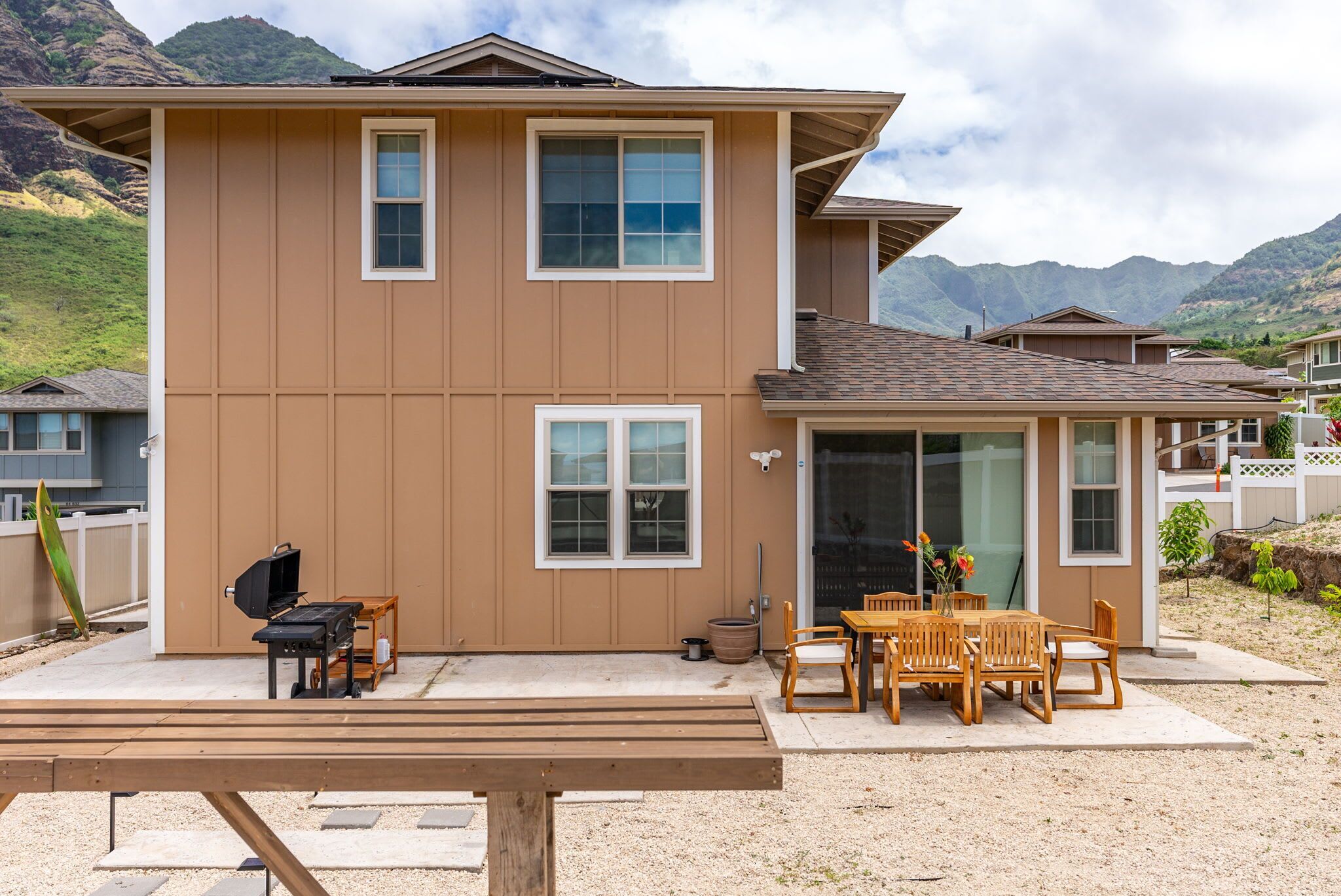 A two-story house with a patio area featuring a grill, wooden chairs, and a table.