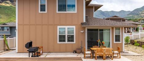 A two-story house with a patio area featuring a grill, wooden chairs, and a table.