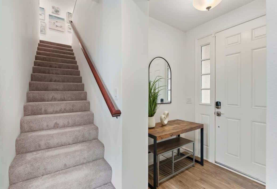 A staircase with carpeted steps and a wooden handrail, leading to a room with a wooden table, a mirror, and white doors.