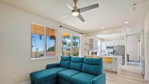 A modern living room with a teal sectional sofa, a ceiling fan, and a kitchen area in the background.