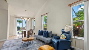 A modern living room with a grey sofa, blue armchair, and a round wooden coffee table. There is a dining area with a wooden table and chairs, and a large window with curtains.