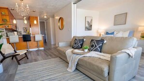 A modern living room with a beige sofa, a chandelier, and a dining area with wooden cabinets and bar stools. - @ Marbella Lane - Vibrant and Refreshing Studio (Honolulu)