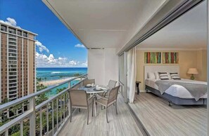 Uma varanda com mesa e cadeiras, uma cama com vista para o oceano e um edifício ao fundo.