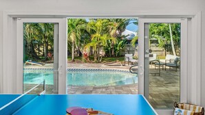 Un espace piscine équipé d’une table de ping-pong, de chaises et offrant une vue sur un jardin agrémenté de palmiers.